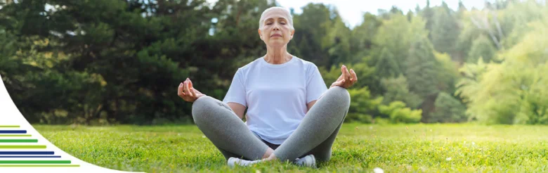 Starší žena sedí na trávě v přírodě při meditaci.
