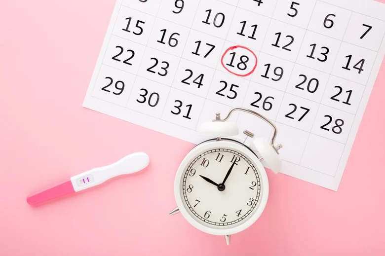 Kalendář s vyznačeným datem, budík a těhotenský test na růžovém pozadí.