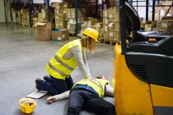 Žena provádí resuscitaci na zraněném kolegovi v skladišti.