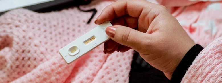 Ruka drží těhotenský test s pozitivním výsledkem na růžovém pozadí.