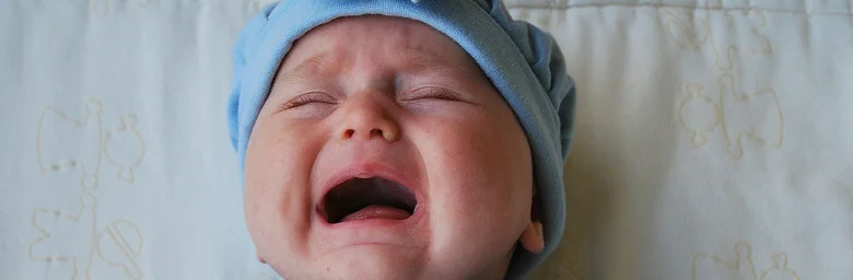 Pláčající dítě v modré čepici na bílém povlečení.