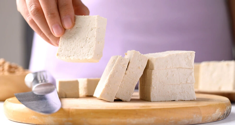 Ruce drží kus tofu nad dřevěnou prkénkem s dalšími kousky.