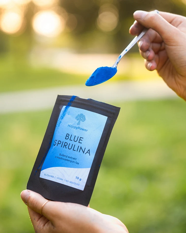 Ruka drží lžičku modré spiruliny nad balením produktu venku.