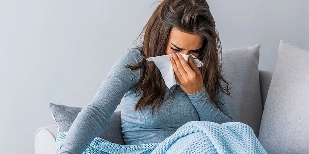 Žena sedí na pohovce s rouškou v ruce při kašli.