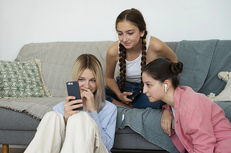 Tři dívky sedí na gauči, jedna drží telefon.