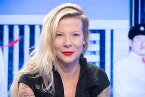 Portrét ženy s blond vlasy v černém oblečení před modrou pozadí.