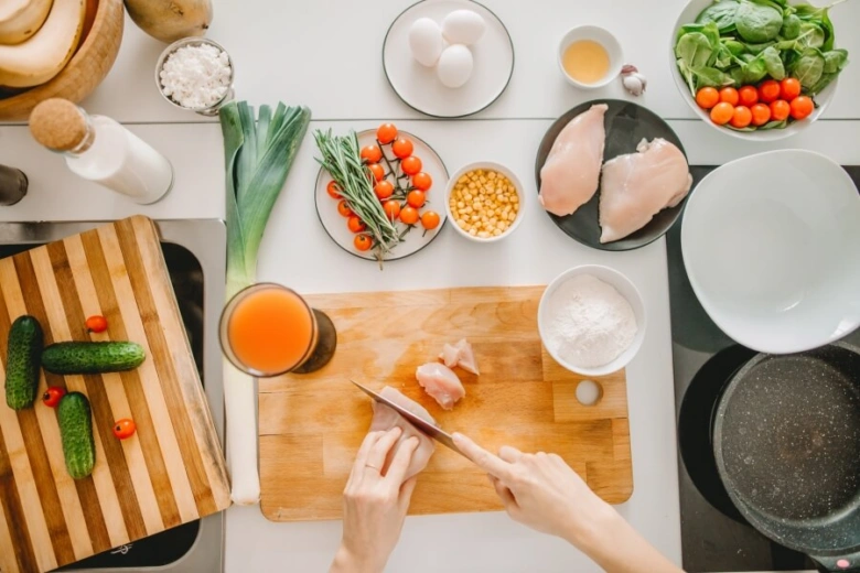 Ruce řezají kuřecí maso na dřevěném prknu s čerstvými zeleninovými ingrediencemi.