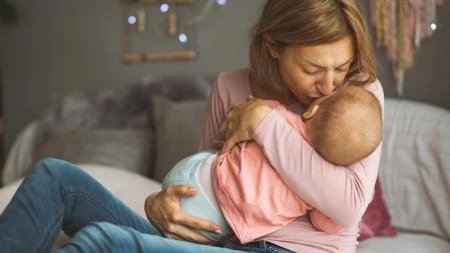 Žena sedí na posteli s novorozencem v náručí.