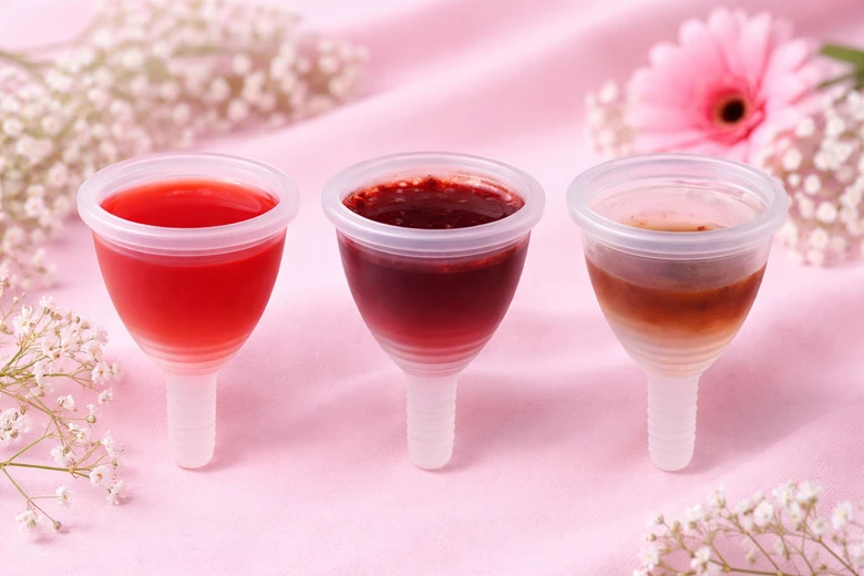 Tři menstruační šálky s červenou až hnědou tekutinou na růžovém pozadí.