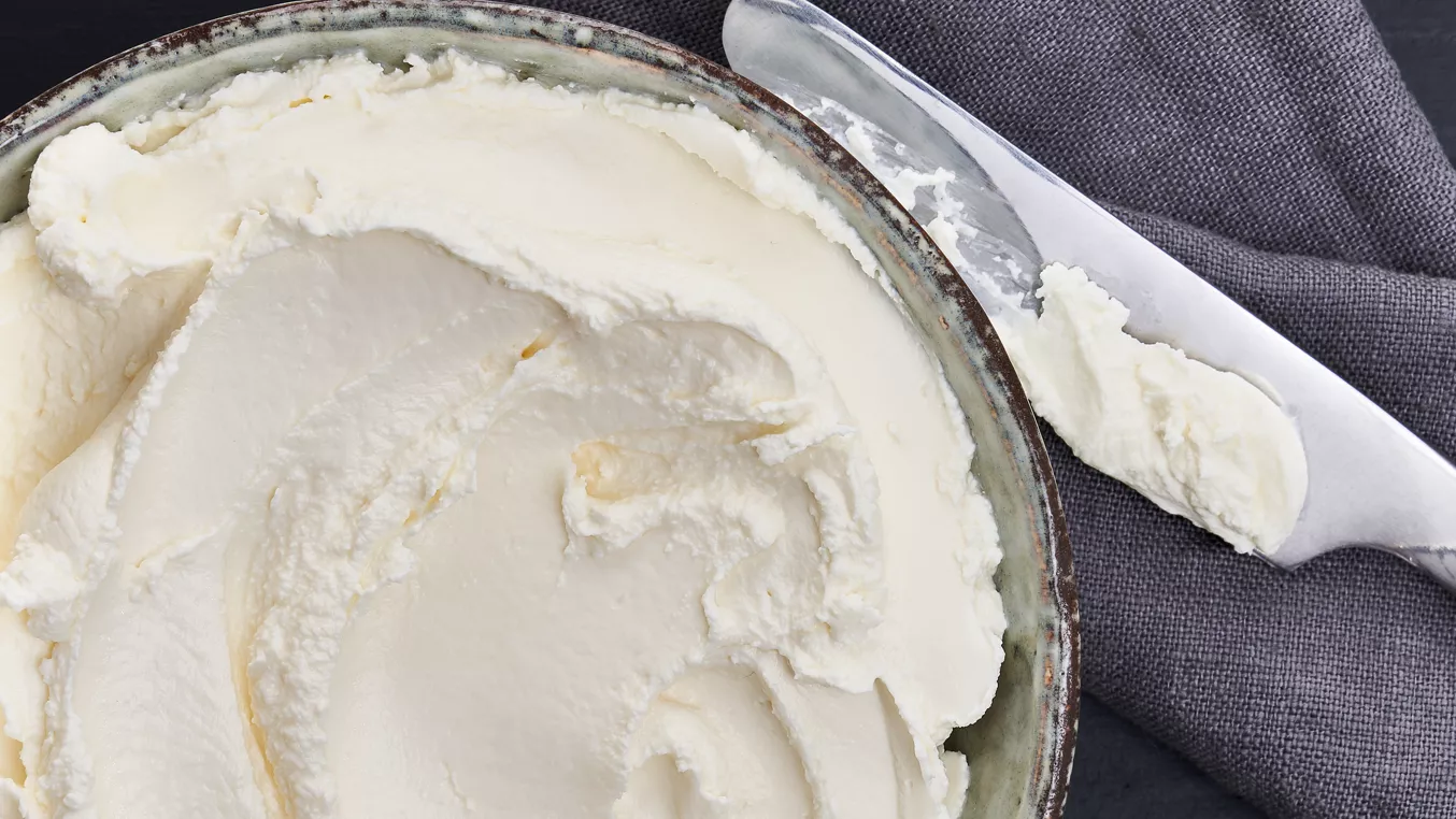 Bílá sýrová ricotta v hrnku vedle lžíce na šedé utěrce.