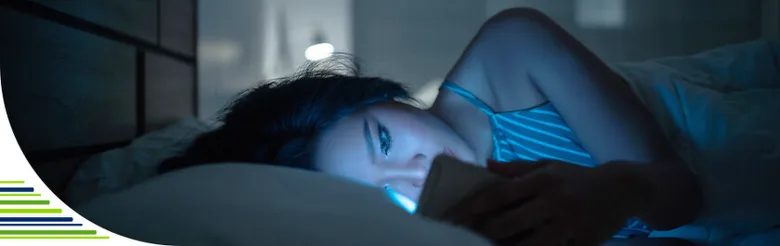 Žena leží v posteli a dívá se na modré světlo ze svého telefonu.