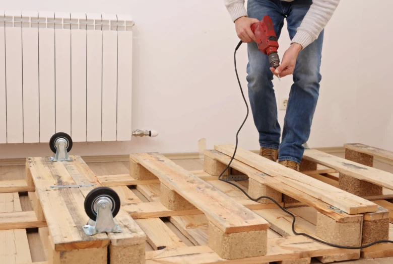 Člověk sestavuje postel z palet pomocí vrtáku