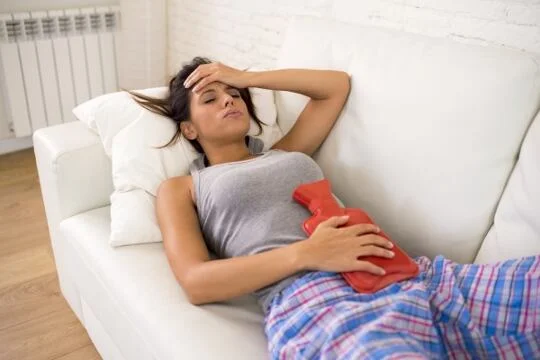 Žena leží na gauči s horkou vodní láhví pro menstruační bolesti.